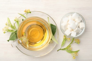 Photo of Fresh linden tea in cup, sugar cubes and flowers on white wooden table, flat lay