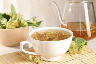 Photo of Fresh linden tea in cup, teapot and flowers on white table, closeup
