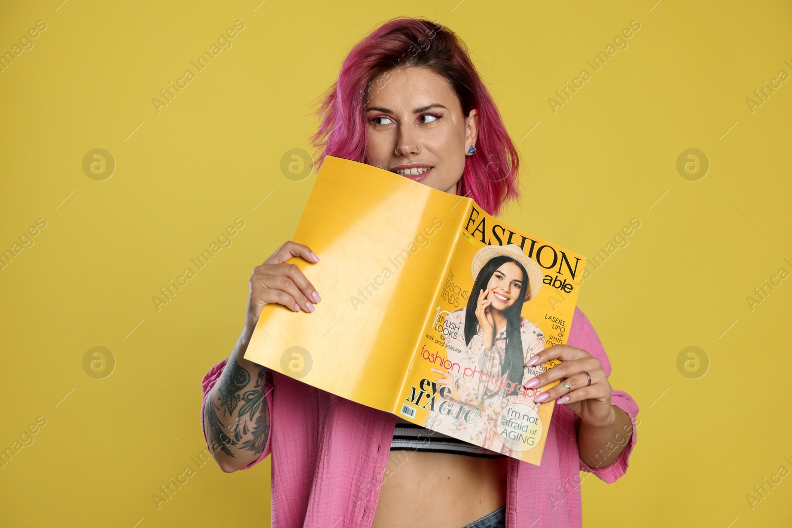 Beautiful woman with pink dyed hair holding fashion magazine on yellow background Photo of Beautiful woman with pink dyed hair holding fashion magazine on yellow background