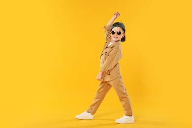 Photo of Happy little boy in sunglasses and baseball hat dancing on orange background. Space for text