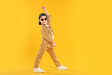 Photo of Happy little boy in sunglasses and baseball hat dancing on orange background. Space for text