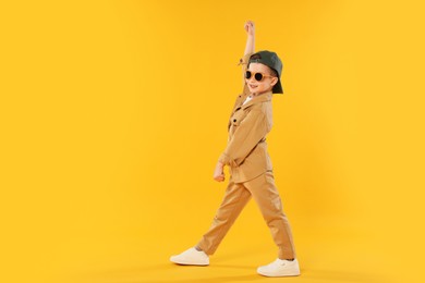 Photo of Happy little boy in sunglasses and baseball hat dancing on orange background. Space for text