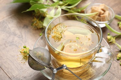 Photo of Fresh linden tea in cup, sugar cubes and flowers on wooden table, closeup