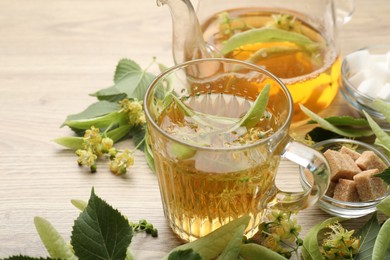 Photo of Fresh linden tea in cup, teapot, sugar cubes and flowers on wooden table, closeup