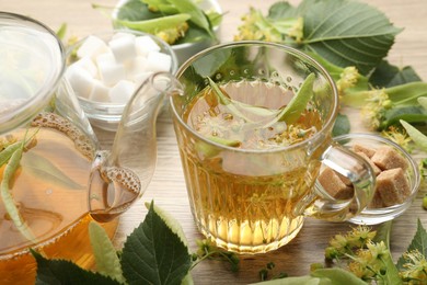 Photo of Fresh linden tea in cup, teapot, sugar cubes and flowers on wooden table, closeup