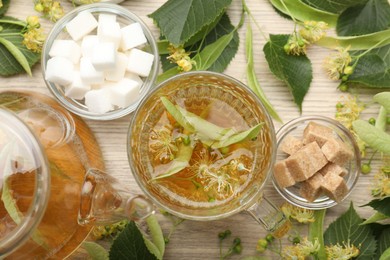 Photo of Fresh linden tea in cup, teapot, sugar cubes and flowers on wooden table, flat lay