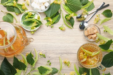 Photo of Frame made of fresh linden tea in cup, teapot, sugar cubes and flowers on wooden table, flat lay. Space for text