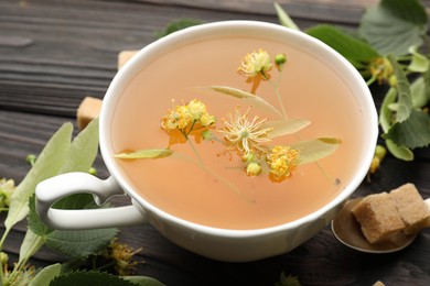 Photo of Fresh linden tea in cup, sugar cubes and flowers on dark wooden table, closeup