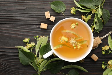 Photo of Fresh linden tea in cup, sugar cubes and flowers on dark wooden table, flat lay