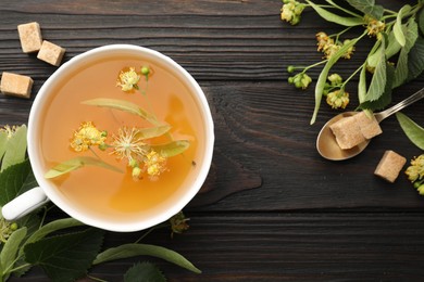 Photo of Fresh linden tea in cup, sugar cubes and flowers on dark wooden table, flat lay. Space for text