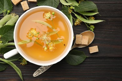 Photo of Fresh linden tea in cup, sugar cubes and flowers on dark wooden table, flat lay