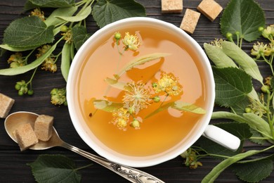 Photo of Fresh linden tea in cup, sugar cubes and flowers on dark wooden table, flat lay