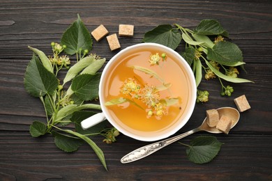 Photo of Fresh linden tea in cup, sugar cubes and flowers on dark wooden table, flat lay
