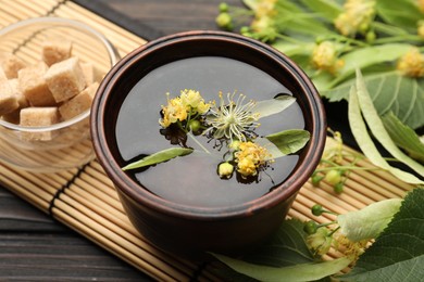 Photo of Fresh linden tea in cup, sugar cubes and flowers on dark wooden table, closeup