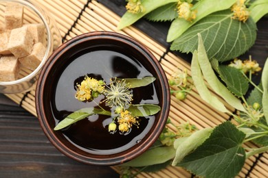 Photo of Fresh linden tea in cup, sugar cubes and flowers on dark wooden table, flat lay