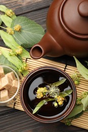 Photo of Fresh linden tea in cup, sugar cubes, teapot and flowers on dark wooden table, flat lay