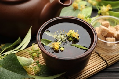 Photo of Fresh linden tea in cup, sugar cubes, teapot and flowers on dark wooden table, closeup
