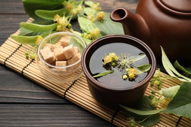 Photo of Fresh linden tea in cup, sugar cubes, teapot and flowers on dark wooden table, closeup