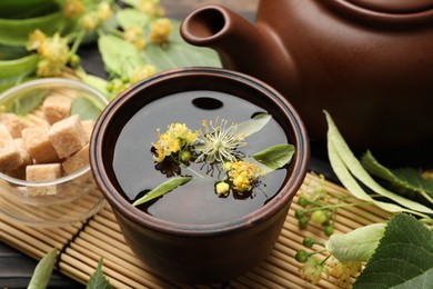 Photo of Fresh linden tea in cup, sugar cubes, teapot and flowers on dark wooden table, closeup