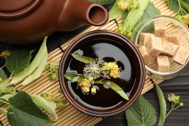 Photo of Fresh linden tea in cup, sugar cubes, teapot and flowers on dark wooden table, flat lay