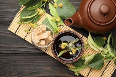 Photo of Fresh linden tea in cup, sugar cubes, teapot and flowers on dark wooden table, flat lay. Space for text