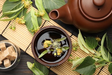 Photo of Fresh linden tea in cup, sugar cubes, teapot and flowers on dark wooden table, flat lay