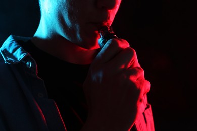 Photo of Man using electronic smoking device in neon lights on black background, closeup