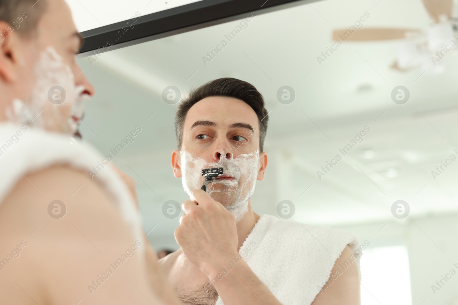 Handsome man shaving with razor near mirror at home, low angle view Photo of Handsome man shaving with razor near mirror at home, low angle view