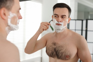Photo of Handsome man shaving with razor near mirror at home