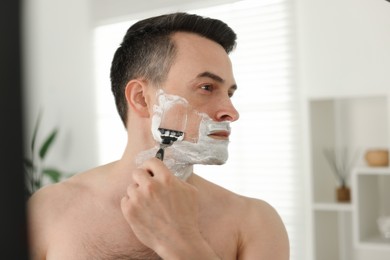Photo of Handsome man shaving with razor near mirror at home