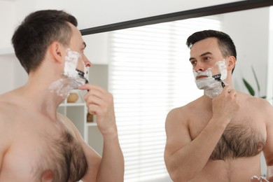 Photo of Handsome man shaving with razor near mirror at home