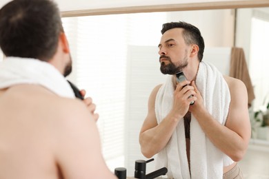 Photo of Handsome man shaving with trimmer near mirror in bathroom