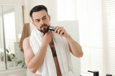 Photo of Handsome man shaving with trimmer near mirror in bathroom