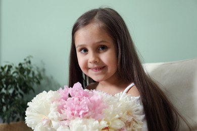 Photo of Cute little girl with bouquet of beautiful peonies in armchair near green wall indoors