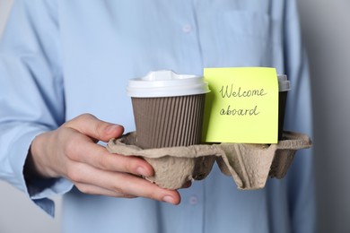 Photo of Welcome Aboard. Woman holding holder with takeaway cups and sticky note on grey background, closeup