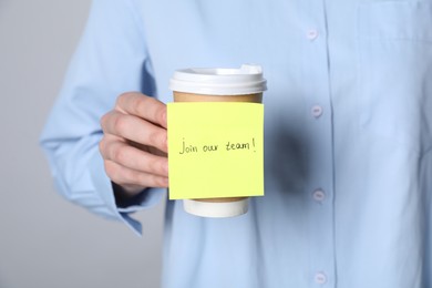 Photo of Join Our Team. Woman holding takeaway cup with sticky note on grey background, closeup