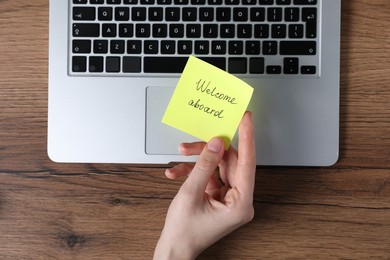 Photo of Woman holding paper note with words Welcome Aboard near laptop at wooden table, top view