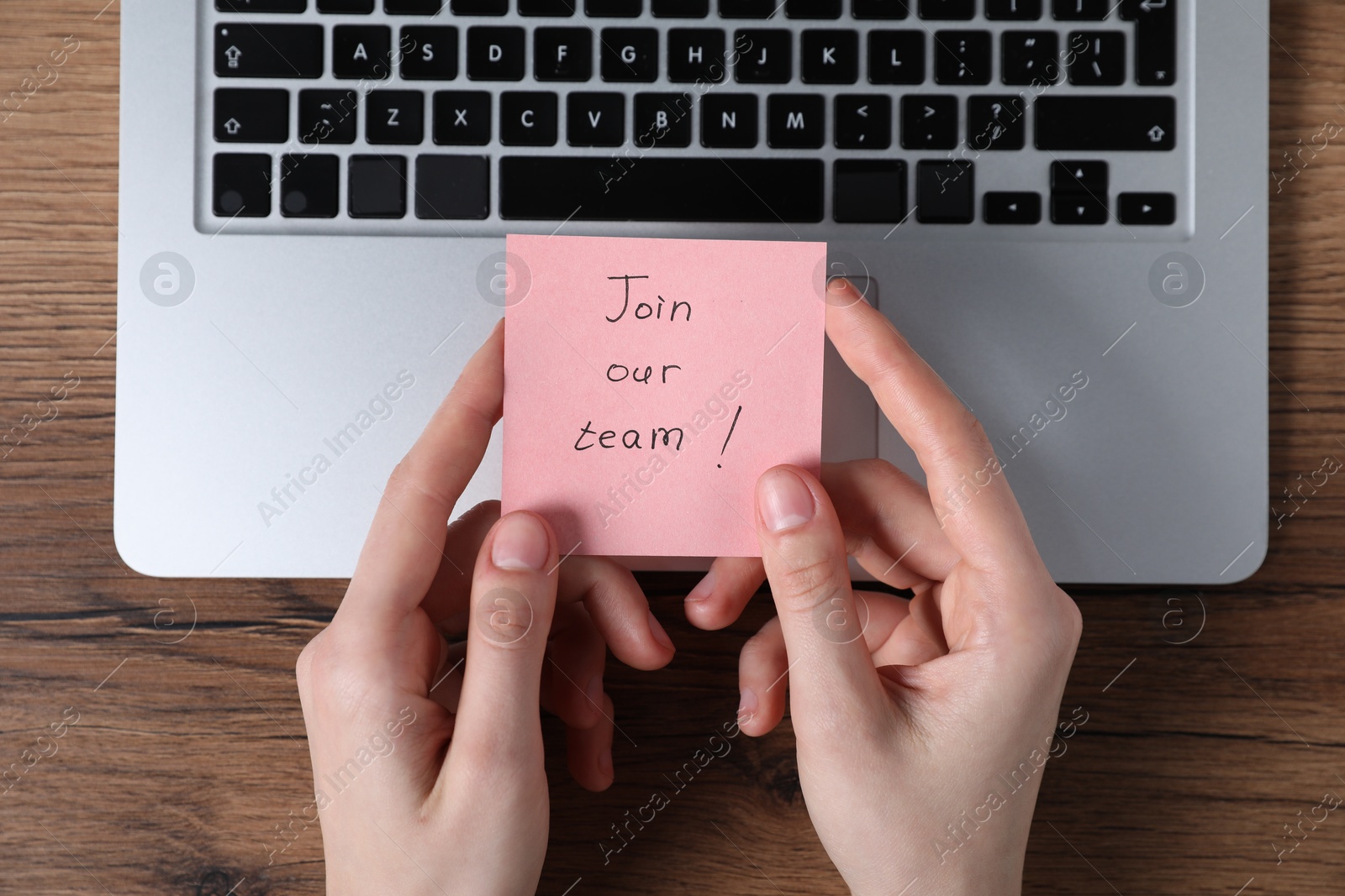 Woman holding paper note with words Join Our Team near laptop at wooden table, top view Photo of Woman holding paper note with words Join Our Team near laptop at wooden table, top view