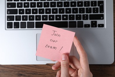 Photo of Woman holding paper note with words Join Our Team near laptop at wooden table, top view