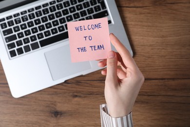 Photo of Woman holding paper note with words Welcome To The Team near laptop at wooden table, top view