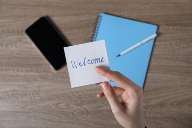 Photo of Woman holding paper note with word Welcome at wooden table, top view