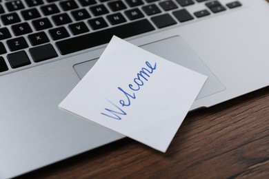 Photo of Paper note with word Welcome and laptop on wooden table, closeup