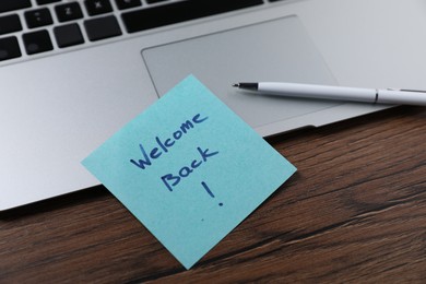 Photo of Paper note with words Welcome Back, laptop and pen on wooden table, closeup