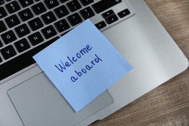 Photo of Paper note with words Welcome Aboard and laptop on wooden table, above view