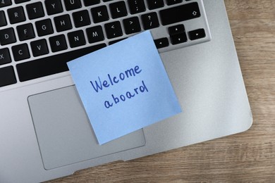 Photo of Paper note with words Welcome Aboard and laptop on wooden table, top view