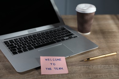 Photo of Paper note with words Welcome To The Team and laptop on wooden table, closeup