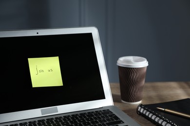 Photo of Sticky note with words Join Us, laptop, notepad and takeaway cup on wooden table against grey background, closeup