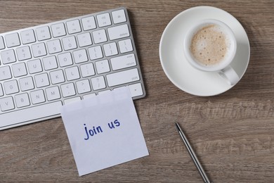 Photo of Paper note with words Join Us, cup of coffee and computer keyboard on wooden table, flat lay