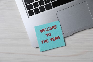 Photo of Paper note with words Welcome To The Team and laptop on light wooden table, top view