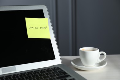 Photo of Sticky note with words Join Our Team, laptop and cup on white wooden table against grey background, closeup